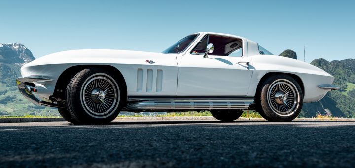 Low side view of a white classic sports coupe on asphalt, with mountains and clear blue sky behind it.